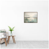 Framed abstract canvas print with soft gray and green tones depicting a hazy horizon, hung on a white wall above a small wooden side table with a green fern in a glass vase.