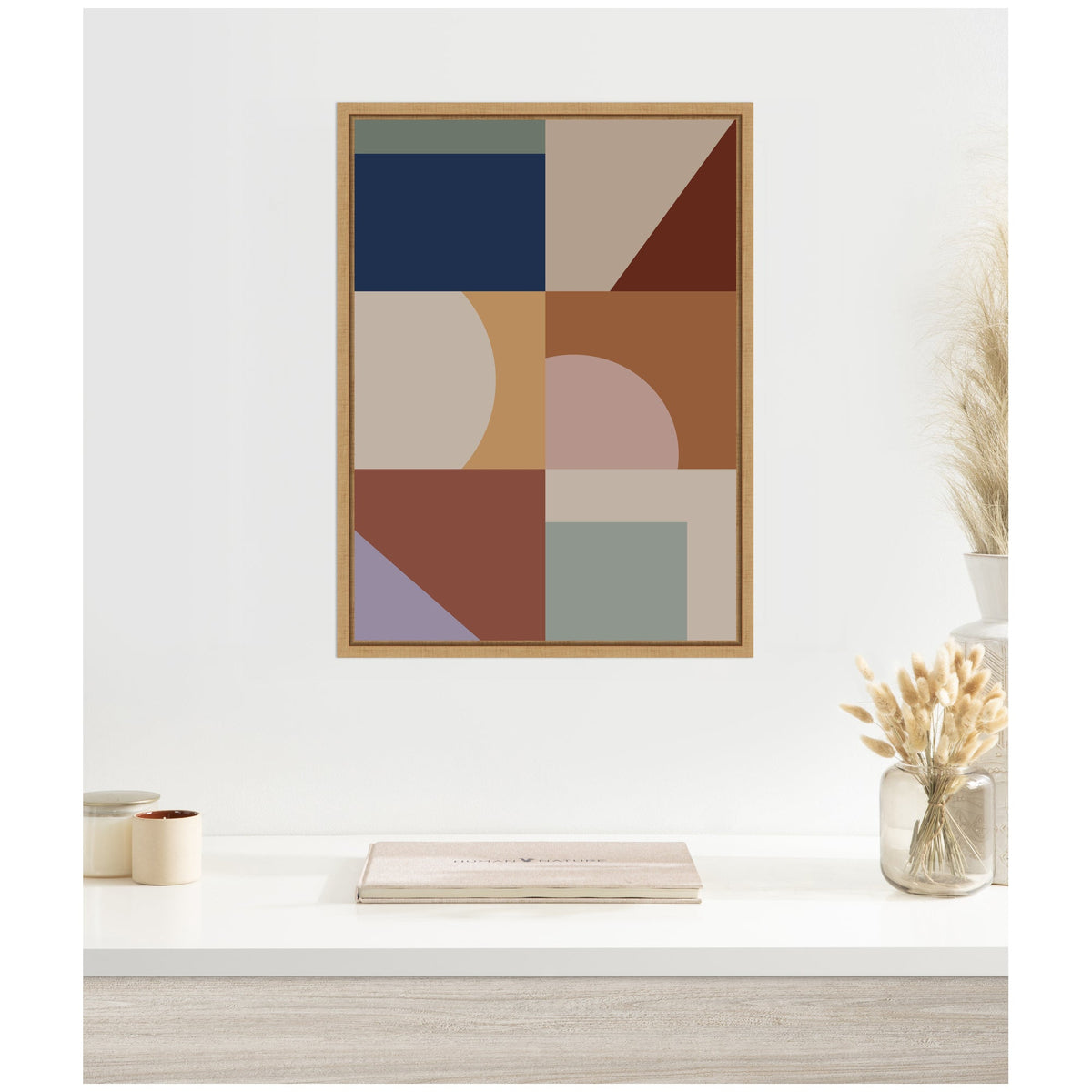 Framed canvas featuring a geometric modern bohemian pattern with blue, beige, rust, mauve, and sage shapes in a wooden frame on a white wall above a minimalist table.