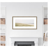 Framed panoramic coastal landscape print with sandy dunes and gentle sea waves in soft, muted green and beige tones above a white mantel surrounded by white shelves with wicker baskets.