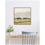 Square framed wall art depicting a muted landscape with rolling hills, green fields, and a cloudy sky, displayed above a light wood desk with a plant and metal decor.