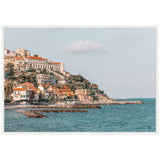 Coastal hillside with pastel-colored buildings and terracotta roofs overlooking turquoise sea under a pale blue sky with single cloud.