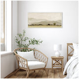 Framed horizontal canvas print featuring soft beige and gray abstract hills with subtle textured strokes, hanging above a woven armchair and wooden nightstand in a bright room.
