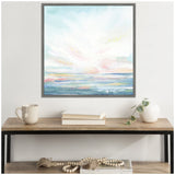 Abstract seascape painting with soft pastel blues, pinks, and whites, depicting a serene sky and reflective water, framed and hung above a wooden table with books and a vase.