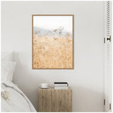 Framed canvas above a wooden bedside table with a cup and two books, featuring a soft-focus photograph of golden wild fennel against a pale sky.
