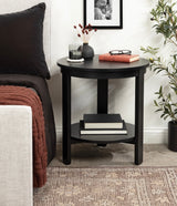 Black round wood side table with lower shelf holding books, a red book and glasses on top, placed beside beige upholstered furniture and a green plant on a patterned rug.