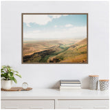 Framed landscape photograph of rolling hills and green pastures under a partly cloudy blue sky, hung above a white dresser with plants, books, and patterned containers.
