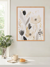 Muted floral abstract print with beige, black, and gray flowers and golden dots in light wood frame on pale wall above white table holding cup, napkin, and pastry.