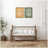 Two framed canvas art pieces with white floral patterns, one on ochre background and the other on olive green, hung above a wooden bench with white cushions in a minimalist room.