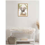 Framed canvas print of an elephant standing in a white clawfoot bathtub with green palm leaves in the background, hung on a light beige wall above a white bench with a fluffy cream throw.