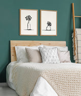Two framed black ink botanical sketches of vases with flowers hang above a bed with a light wood headboard against a teal wall, with a textured cream throw and geometric pillows.