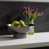 Matte gray ribbed metal bowl with green apples inside, placed on a white surface with a clear jar of purple tulips in the background against a dark paneled wall.