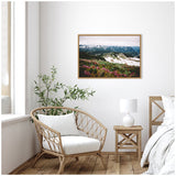 Framed canvas showing a snow-covered mountain landscape with green and pink flora in the foreground, displayed on a white wall above a rattan chair and wooden side table inside a bright room.