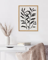 Black botanical silhouette of elongated leaves on white background in natural wood frame above white desk with books and beige knitted throw.