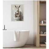 Framed canvas showing a cute donkey sitting on a toilet holding a roll of toilet paper, displayed on a white wall above a freestanding bathtub with a white towel.