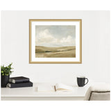Framed landscape painting of soft, muted earth tones depicting rolling hills under a cloudy sky, hanging above a white desk with books, plant, coffee cup.