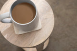 White ceramic mug with coffee on a round light wood table with subtle grain, viewed from above against a neutral background.