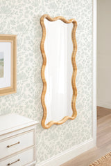 Tall rectangular mirror with a scalloped frame made from natural light rattan hanging on a pale floral-patterned wall beside a white dresser.