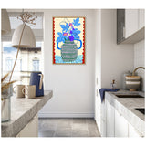 Framed canvas art of a blue and white patterned pot with bright blue and purple fig leaves, hanging on a white kitchen wall with stone countertops and a blue pitcher nearby.