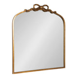 Arched mirror with a thin gold frame topped by an ornate ribbon-shaped bow detail in the same gold finish, reflecting a plain background.