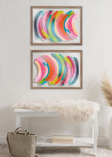 Two framed abstract prints with overlapping crescent shapes in pink, green, blue, and orange, displayed vertically on a neutral wall above a white bench with a fur throw.