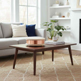 Mid-century modern brown wood coffee table with tapered legs, topped with a round copper and light wood decorative box and a stack of books, in a bright living room.