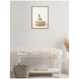 Framed canvas artwork of a cheetah sitting inside a beige terrazzo bathtub with scattered spots, hung above a white bench draped with a fluffy cream fur throw in a minimal room.