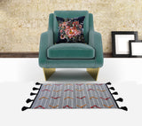 Teal velvet armchair with gold metal legs, featuring a dark velvet pillow adorned with a multicolored floral design, set against a textured beige stone wall with two unframed artworks leaning on the floor nearby.