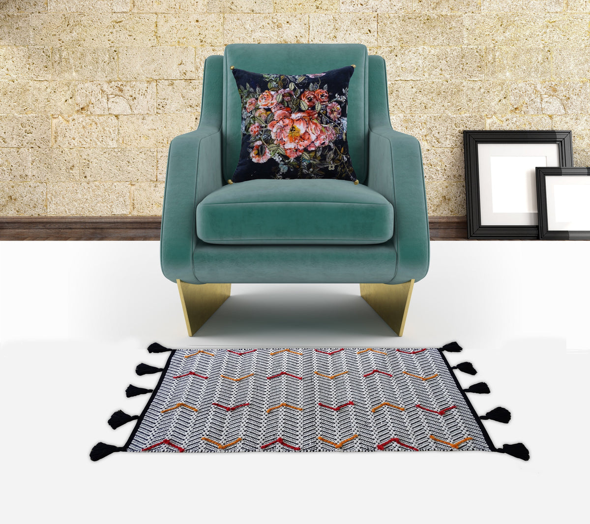 Teal velvet armchair with gold metal legs, featuring a dark velvet pillow adorned with a multicolored floral design, set against a textured beige stone wall with two unframed artworks leaning on the floor nearby.