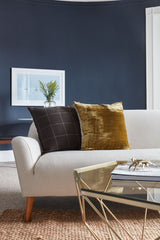 Beige sofa with two decorative pillows, one dark brown with a grid pattern and one golden brown velvet, against a deep blue curved wall with framed artwork.