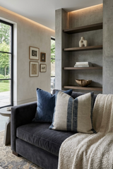 Dark gray velvet sofa with an indigo blue pillow and a beige pillow featuring vertical indigo stripes, next to a textured beige throw blanket under built-in concrete shelves.