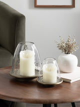 Two hammered glass cloche candle holders with white pillar candles inside, sitting on round brass bases on a wooden coffee table beside a white ceramic vase with dried flowers.