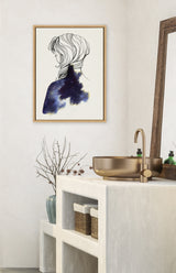 Framed canvas artwork showing a black and white line drawing of a woman with hair flowing into a dark blue and black galaxy-like pattern on her shoulder, hanging on a pale wall above a beige textured vanity with a round gold sink,