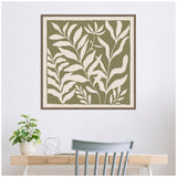 Square framed canvas with minimalist botanical design of cream leaves on olive green background above a light wood desk with potted plant and gray chair