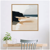 Abstract framed canvas of a coastal landscape with beige sand, dark cliffs, and muted blue water under a pale gray sky, hanging above a wooden desk with a plant and chair.