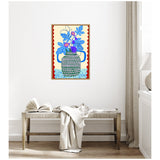 Colorful framed canvas print of a stylized pot with blue and purple fig leaves and fruits on a pale yellow background with a red wavy border, hung above a rustic wooden bench with a white throw and open book.