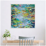 Framed canvas painting of a pond with multicolored water lilies and green foliage, mounted on a white wall above a wooden desk with a potted plant and gray chair.