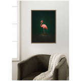 Framed wall art print of a pink flamingo standing against a rich green background, hung above a dark olive green armchair draped with a beige knit blanket.