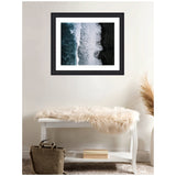 Framed aerial photo of ocean waves crashing onto a black volcanic beach with white foam, displayed above a white bench with a fur throw and a brown bag beneath it.