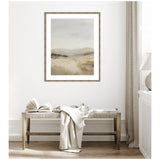 Framed abstract landscape painting with soft beige and gray tones hanging above a wooden bench with a light fabric draped over it in a minimalist room.