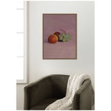 Framed still life painting of a peach, dark plum, and green grapes on a soft purple background, hung above a dark olive armchair with a white knit throw.