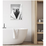 Black and white framed photo of a capybara wrapped in a towel, hanging above a white freestanding bathtub with a towel draped over it in a minimal wooden bathroom.