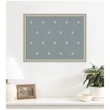 Framed blue botanical block print with a repeating pattern of small white stylized leaves, hanging above a white surface with a lamp, photo frame, bowl, and potted plant.