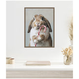 Framed canvas artwork of a brown and white bunny holding a red and white striped candy cane against a soft gray background, hanging above a light wooden desk with beige decor.
