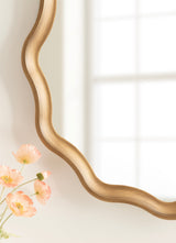 Close-up of a scalloped round mirror with a natural wood frame in light brown tones, reflecting soft window light and partially showing peach-colored flowers nearby.