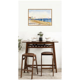 Framed canvas painting of a beach scene with blue sea and sandy shore hanging above a dark wood bar table with two matching stools and a small potted plant at center.