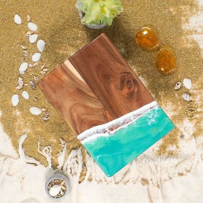Wooden resin charcuterie board with turquoise resin on a sandy background with seashells and stones.