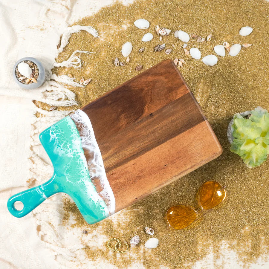 Wooden resin charcuterie board with teal handle on a sandy background with seashells and ocean-themed decor.
