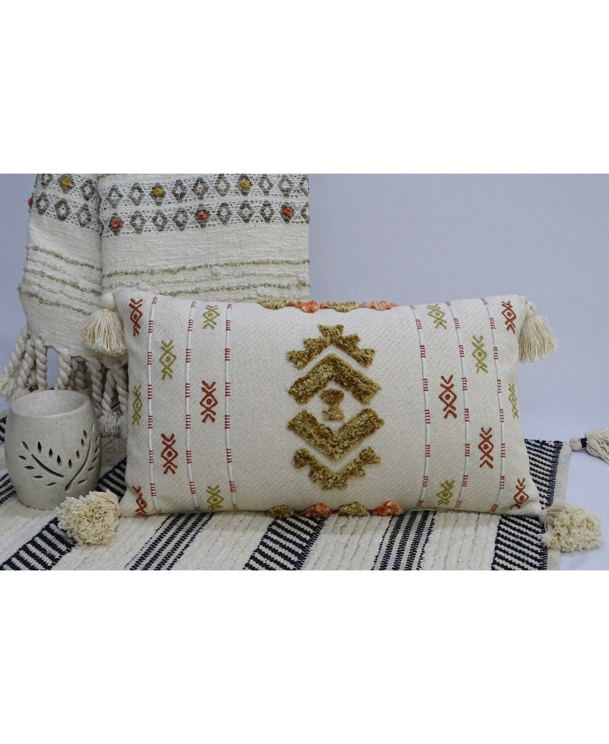 Rectangular cream throw pillow with tribal-style embroidery in mustard, rust, and olive green, featuring large central geometric motifs and tasseled corners on a striped rug.