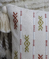 Close-up of a cream-colored throw pillow with vertical red and olive embroidery patterns and a large cream tassel at the corner against a textured cream background.
