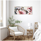 Horizontal floral canvas art with large pink and white flowers hangs on white wall above a wicker chair with white cushion and wooden side table with white lamp in bright room.
